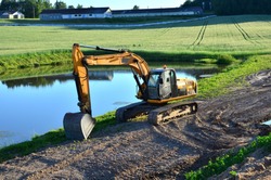 Excavator by the river against the green field of the countryside during the construction of the highway - Image