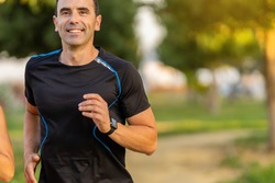 Joyful adult man working out in the park and jogging. He is smiling and looking at front.