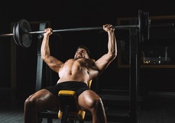 Adult caucasian male shirtless doing bench presses with weights in a bar in a dark gymnasium
