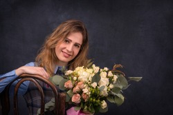 Close up portrait of the young woman with a bouquet of roses. She sits on the Vienna chair and smiles. A photo in retrostyle against a dark background in studio.