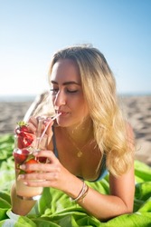 Blonde woman drinking cocktail. Woman holding glass with bright cold beverage. Lying on blanket at beach. Party, outdoor activity, beverages concept