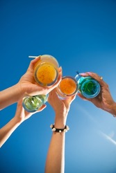 Close-up of friends clinking glasses. Multicolored fancy drinks in glasses, women clinking glasses. Party, outdoor activity, beverages concept