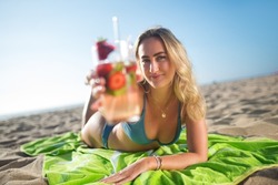 Blonde young woman drinking cocktail. Woman holding glass with bright cold beverage. Lying on blanket, looking at camera. Party, outdoor activity, beverages concept