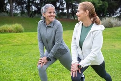 Cheerful senior friends exercising in park. Women in sportive clothes stretching on cloudy day. Sport, friendship concept