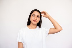 Portrait of thoughtful young woman scratching head over white background. Caucasian lady wearing white T-shirt looking away with pensive expression. Planning or dreaming concept