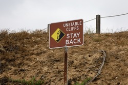 An unstable cliffs stay back sign with pictograph of rocks falling on top of a person.