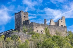 Ruined Brandenbourg Castle on a promontory surrounded by small green trees, towers and stone walls, blue sky and few white clouds in the background, sunny summer day in Luxembourg