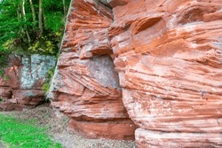 Red sandstone rocky slope, irregular texture, mold, white spots and linear erosions caused by passage of time, trees with green foliage in background, sunny day around Lake Stausee Bitburg, Germany