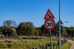 Traffic sign indicating: maximum speed 30 and men working, work zone in road construction and repair, Dutch countryside with trees in background, sunny summer day in Meers, Elsloo, Netherlands