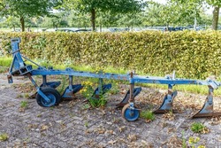 Blue metal agricultural tractor plow in a farm yard, rusty and abandoned, cars parked in parking area in background, bushes and trees, sunny day in Sweikhuizen, South Limburg, Netherlands