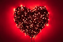 Shape of heart folded from tangled wires of a garland with red light bulbs on red background in the dark with a vignetting effect