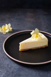 Classic New York cheesecake with fresh vanilla flower on a dark concrete background, side view. A piece of Vanilla cheesecake on a black plate. Confectionery menu, recipe. Close up, Vertical