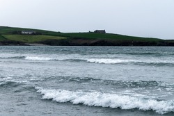 Sea waves at high tide. A building on a hill on the seashore. Gray gloomy sky, building on green grass field near sea. Seaside landscape, white foam on the waves.