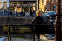 Backside of a old man on wooden bench with blurred view along the canal, The senior guy sitting and relaxing, Hobby and leisure activity of retirement age, Amsterdam capital city of the Netherlands.