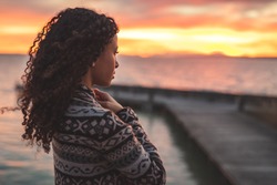 Young African American woman is standing on the promenade at the lake, looking thoughtfully towards the water and the setting sun. The girl in a vest is in a thoughtful mood