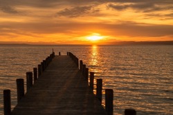 Atmospheric photo of the sunset at Lake Neusiedl with a wooden footbridge running from the picture in the foreground to the left. The wooden poles cast a shadow on the jetty. Sun is reflected in water