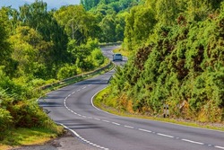 A car drives on a road. A narrow, winding road in Scotland along Loch Ness. Trees and bushes next to the road in summer in sunshine. Traffic signs and guard rails