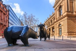 Place in front of the entrance to the Frankfurt Stock Exchange. Bull and bear as a symbol figure. Commercial buildings with a brown facade in the sunshine and blue sky with clouds in spring