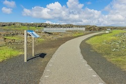 Pathway with a sign pointing to a metal bridge on the Reykjanes Peninsula of Iceland. Lava rocks and sand with green grass between the American and Eurasian tectonic plates