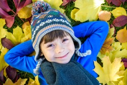 Little kid boy lying in autumn leaves in blue pullover. Happy child having fun in autumn park on warm day. Cute school boy smiling and laughing