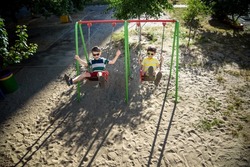Two little kid boys having fun with swing on outdoor playground. Children, best friends and siblings swinging on warm sunny spring or autumn day. Active leisure with kids. Casual boy fashion.