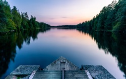 A calm and silent evening at sunset at a small forest lake in Sweden. In the foreground there is a wooden bridge. The lake is surrounded by trees that are reflected in the water