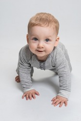 Cute baby crawling on white studio background, looking at you, smiling. Child is dressed in gray jumpsuit. Blond caucasian boy with blue eyes smiles cheerfully. Eye contact. Advertising baby concept.