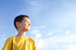 Child standing in profile blue sky with clouds on background. Boy 8 years old brunette, smiling face. Kid in yellow T-shirt. Concept of childhood, growing up, peace, dreams, children. Sunny day.