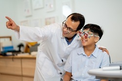 Ophthalmologist or optical staff point to the direction for viewing of Indian boy who wear special glasses to test his eye during process of eyecare in the shop.