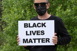 Protests broke out across the United States over the death of George Floyd. A man in a medical black mask and black glasses holds a white banner with the text BLACK LIVES MATTER.