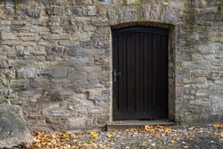 Antique wooden door with an arch in a stone wall. Yellow autumn leaves on the sidewalk.