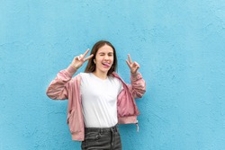 Portrait of funny caucasian teen girl in blank white t-shirt and pastel pink bomber jacket against blue city wall solid background. 