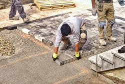 Professional landscape workers paving driveway interlock patio with stone brick at residential construction site. Contractor wearing safety protective cloth on  heavy installation yard work project.