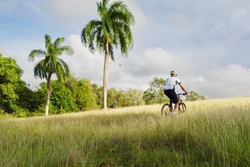 Rear view. Man riding uphill on mountain bike in grass. Tropical environment.
