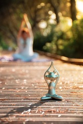Backlight of a yoga frog figure at foreground and a defocused woman at background both in lotus meditating pose with hands above head, at sunset