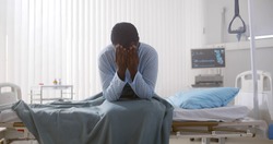 Portrait of afro-american sick patient sitting on hospital bed and rubbing face feeling unwell. Tired and exhausted sick black man sitting in hospital ward
