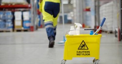 Cropped shot of janitor with bucket washing floor in warehouse focus on yellow cart with industrial worker cleaning floor with mop on storehouse background