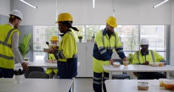 Construction company workers having takeaway lunch in modern office. Multiethnic team of engineers in safety uniform and hardhat taking food from table in office