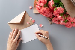 woman writing greeting card on gray table with bouquet of pink roses. Writing letter concept. top view.
