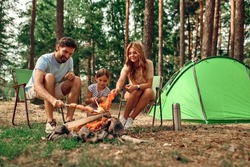 Happy family with a child on a picnic sit by the fire near the tent and grill a barbecue in a pine forest. Camping, recreation, hiking.