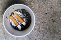 Two cigarette butts and their ashes at the bottom of white paper cup, with hard floor background. Concept of smoking addiction, smoking area