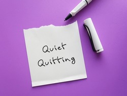 On purple background, pen writing on paper note QUIET QUITTING, when employees not engaged or taking job seriously, do minimum required but focus on job outside office