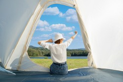 Happy Asian woman sitting with stretching her arms in a camping tent while admiring beautiful calm lake with dense woods and clear blue sky with white clouds in front of her during a warm sunny day.