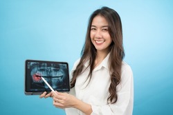 Front view half-body image of a smiling Asian woman standing holding a tablet showing panoramic dental x-rays and using a pen pointing to a red area of problem tooth isolated on turquoise background.