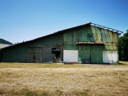 Front view of a large old rusted metal barn with a partially damaged roof, built on the grass floor in rural farms surrounded by  green trees with a clear blue sky in the background on a bright day.