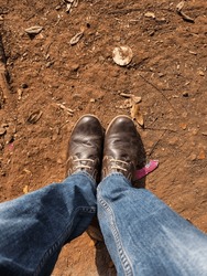Top angle view of men leg in Blue color jeans with old damaged brown color shoes on stony clay ground on background. Picture captured under bright sunlight at Kolhapur, Maharashtra, India.