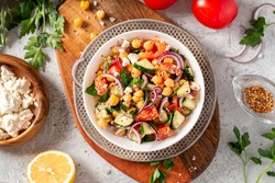 Chickpea salad with tomatoes, cucumber, feta cheese, parsley, onions and lemon in a plate on a gray background top view, selective focus. Healthy vegetarian food, oriental and Mediterranean cuisine.