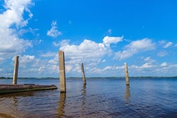 Concrete pillars, the remains of a pier at the river bank. In the distance, a wide river expanse and snow-white clouds in the blue sky open up. The opposite shore is visible