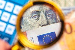 A person with a magnifying glass looking for money. Dollar and Euro together being observed in a photo with a blurred background. International business and world economy concepts.
