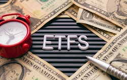 The initials ETFS (Exchange Traded Funds) in a lettered frame with US dollar banknotes, a pen and a red alarm clock in the composition.
